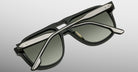 The Jacques Marie Mage Leonard sunglasses, featuring black frames, green-tinted lenses, and silver metal accents with textured silver arms, are shown folded on a light gray surface.