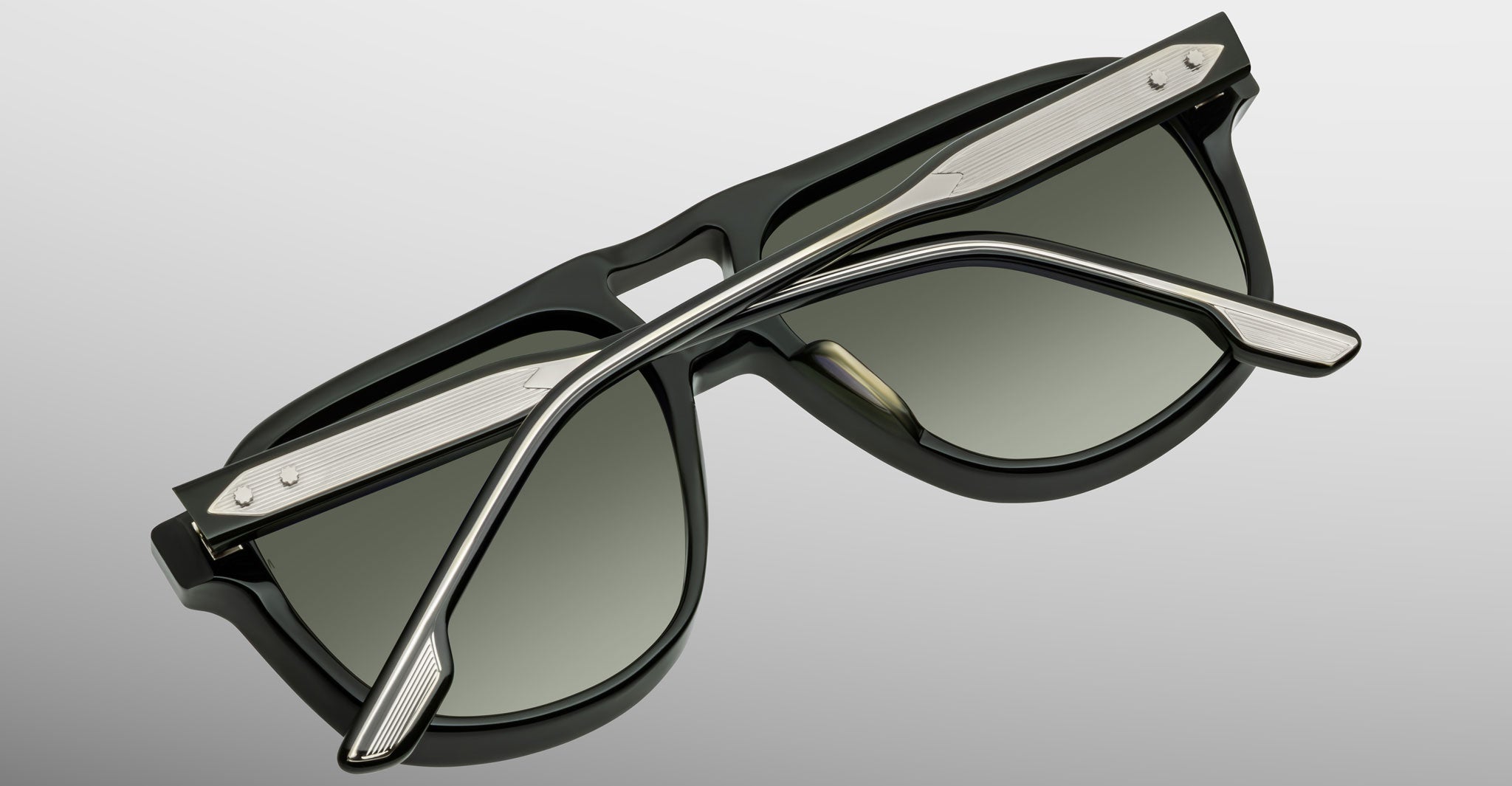 The Jacques Marie Mage Leonard sunglasses, featuring black frames, green-tinted lenses, and silver metal accents with textured silver arms, are shown folded on a light gray surface.