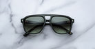 A pair of Jacques Marie Mage Leonard black-framed sunglasses with dark tinted lenses rests on a white marble surface, displayed from above with the arms open and lenses facing up.