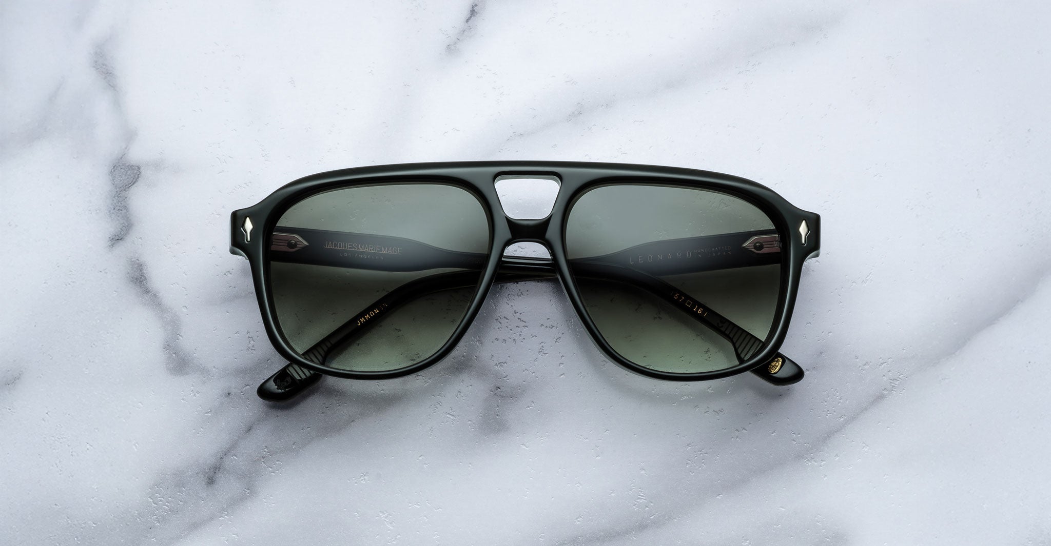 A pair of Jacques Marie Mage Leonard black-framed sunglasses with dark tinted lenses rests on a white marble surface, displayed from above with the arms open and lenses facing up.