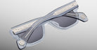 Jacques Marie Mage Lupine sunglasses, shown folded, feature clear frames with dark lenses. The temples include silver-tone metal detailing with engraved patterns and round emblems near the hinges.