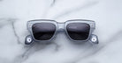 A pair of Jacques Marie Mage Lupine rectangular gray transparent sunglasses with dark lenses rests on a white marble surface.