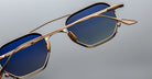 Close-up of Jacques Marie Mage Marbot sunglasses featuring gold metal frames and blue gradient lenses, set against a plain gray background.