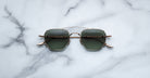 A pair of Jacques Marie Mage Marbot gold-framed sunglasses with green lenses rests on a white marble surface streaked with gray veining.