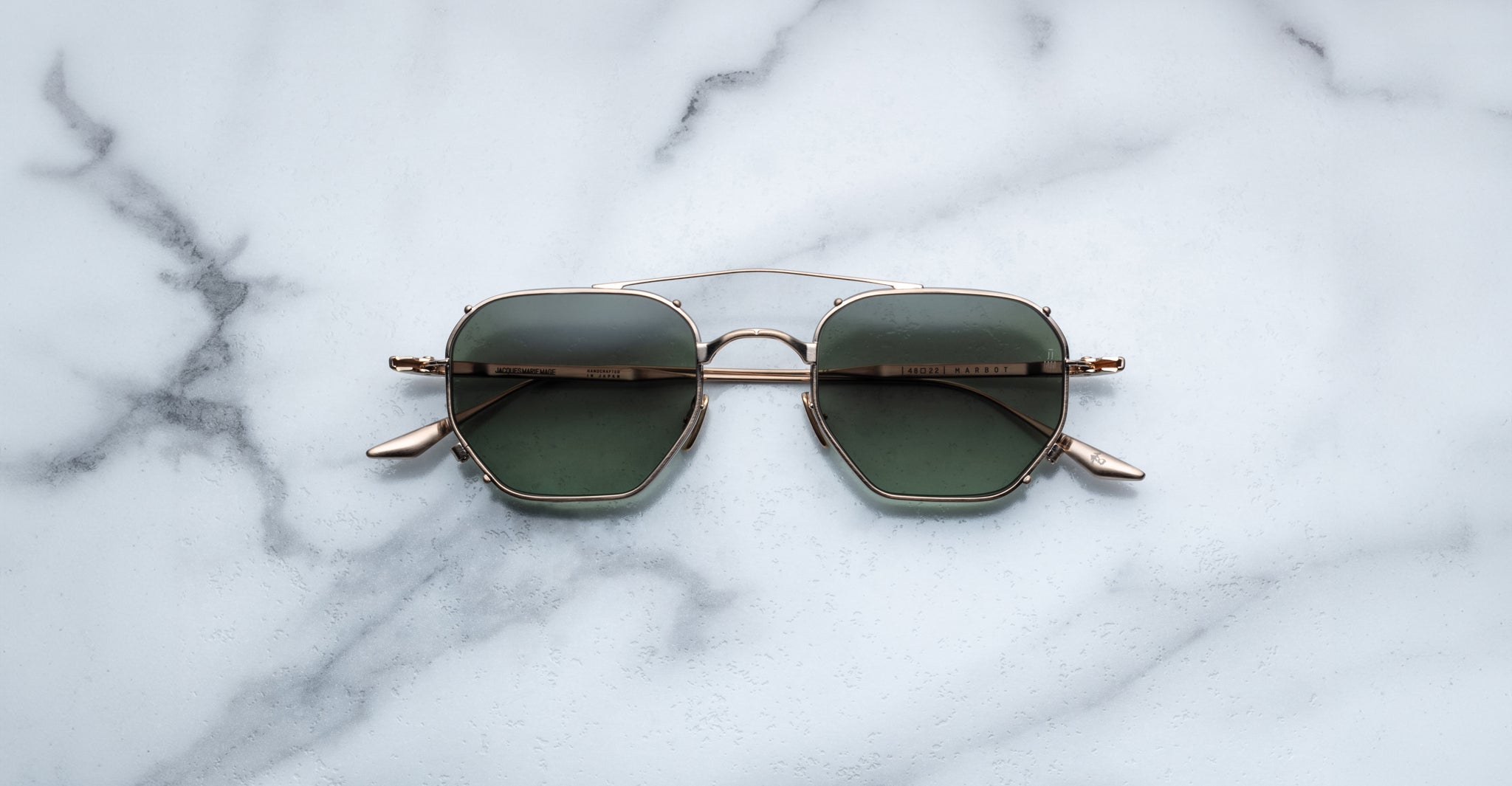 A pair of Jacques Marie Mage Marbot gold-framed sunglasses with green lenses rests on a white marble surface streaked with gray veining.