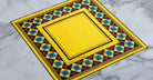 Jacques Marie Mage Maxime yellow square scarf with red, blue, and black geometric border, top view.