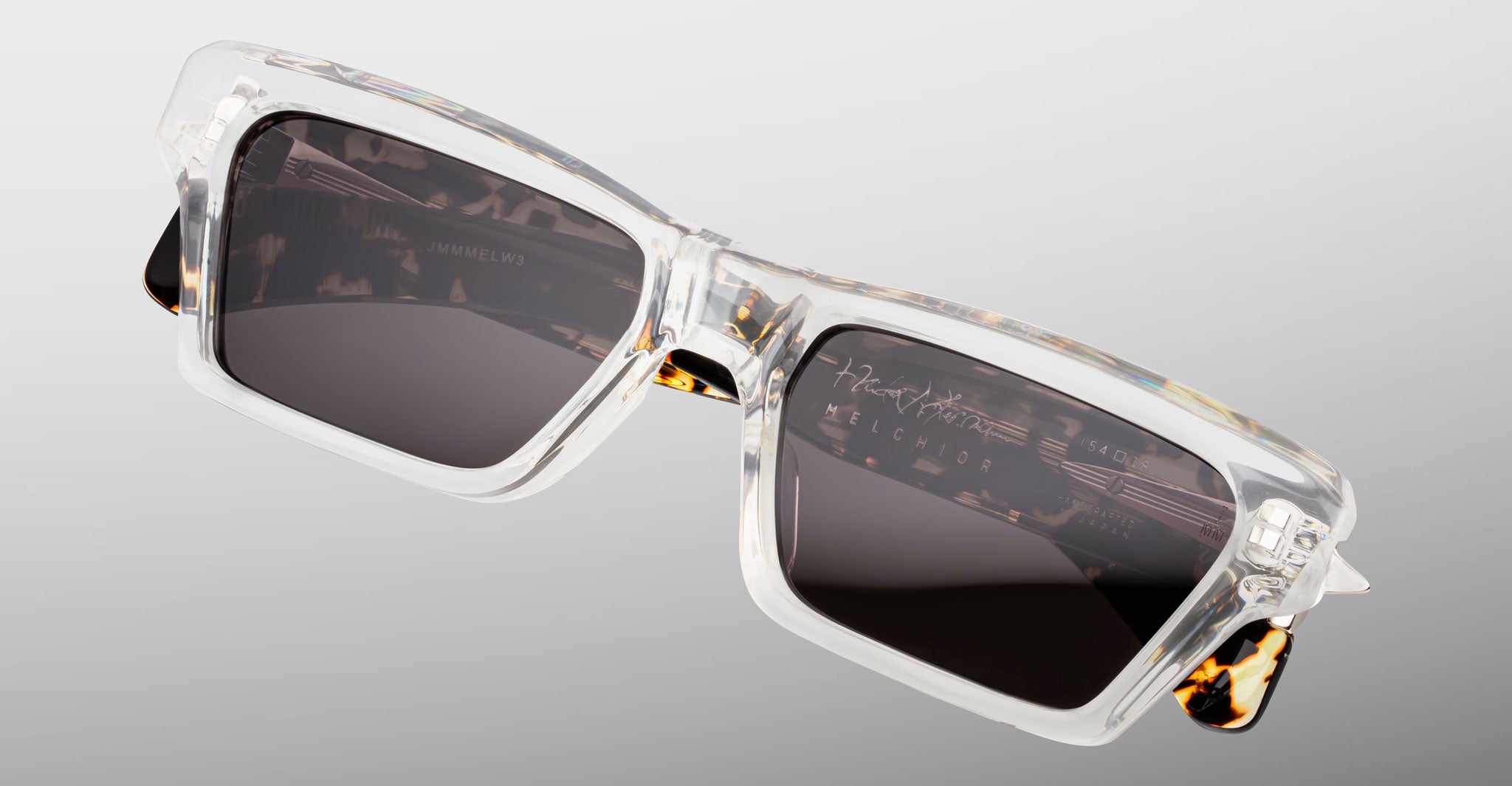 Jacques Marie Mage Melchior sunglasses with clear frames, dark square lenses, and tortoiseshell arms shown from a front angle.