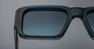 Close-up of the top half of Jacques Marie Mage Miglia rectangular sunglasses in teal with gradient tinted lenses, set against a plain, light gray background.