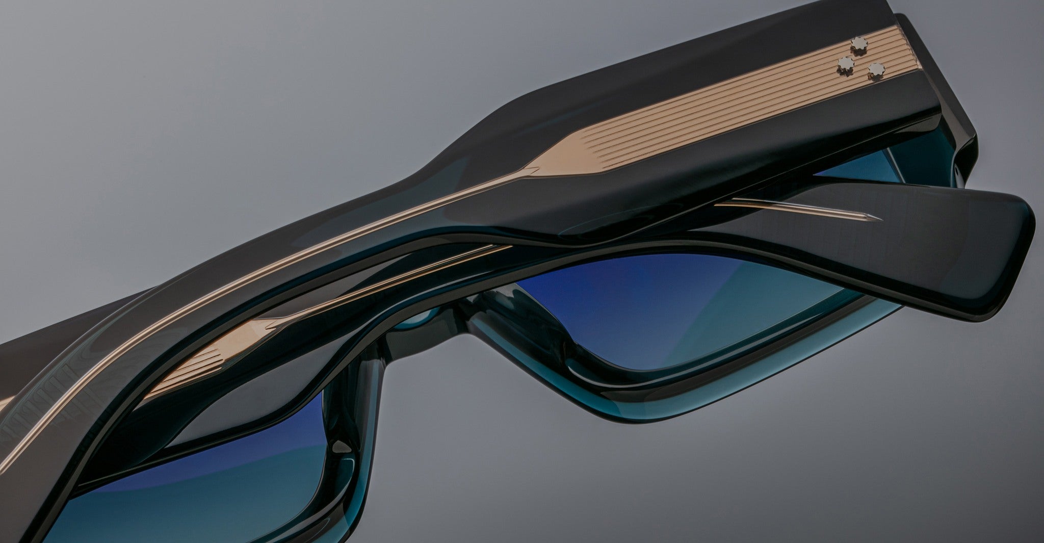 A close-up of two pairs of Jacques Marie Mage Miglia sunglasses with dark frames and blue-tinted lenses, stacked together, highlighting their metallic accents and detailed arms.