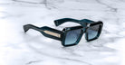The Jacques Marie Mage Miglia sunglasses feature dark, thick rectangular frames and metallic accents on the arms, elegantly displayed on a white marble surface.