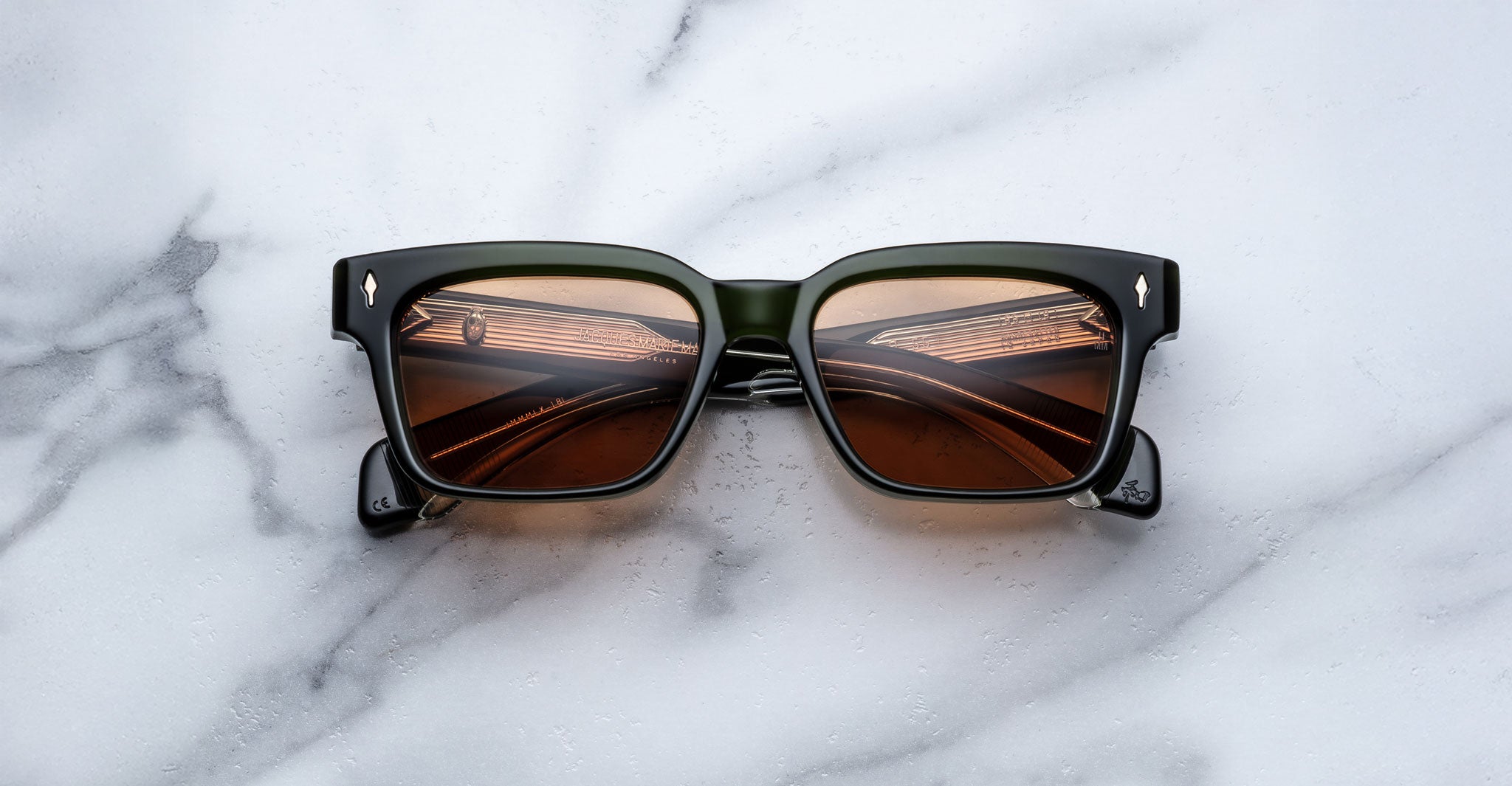 A pair of Jacques Marie Mage Molino 55 black square-framed sunglasses with brown lenses rests on a white and gray marble surface, the lenses reflecting lights and a ceiling structure.
