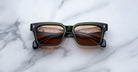 A pair of Jacques Marie Mage Molino 55 black square-framed sunglasses with brown lenses rests on a white and gray marble surface, the lenses reflecting lights and a ceiling structure.