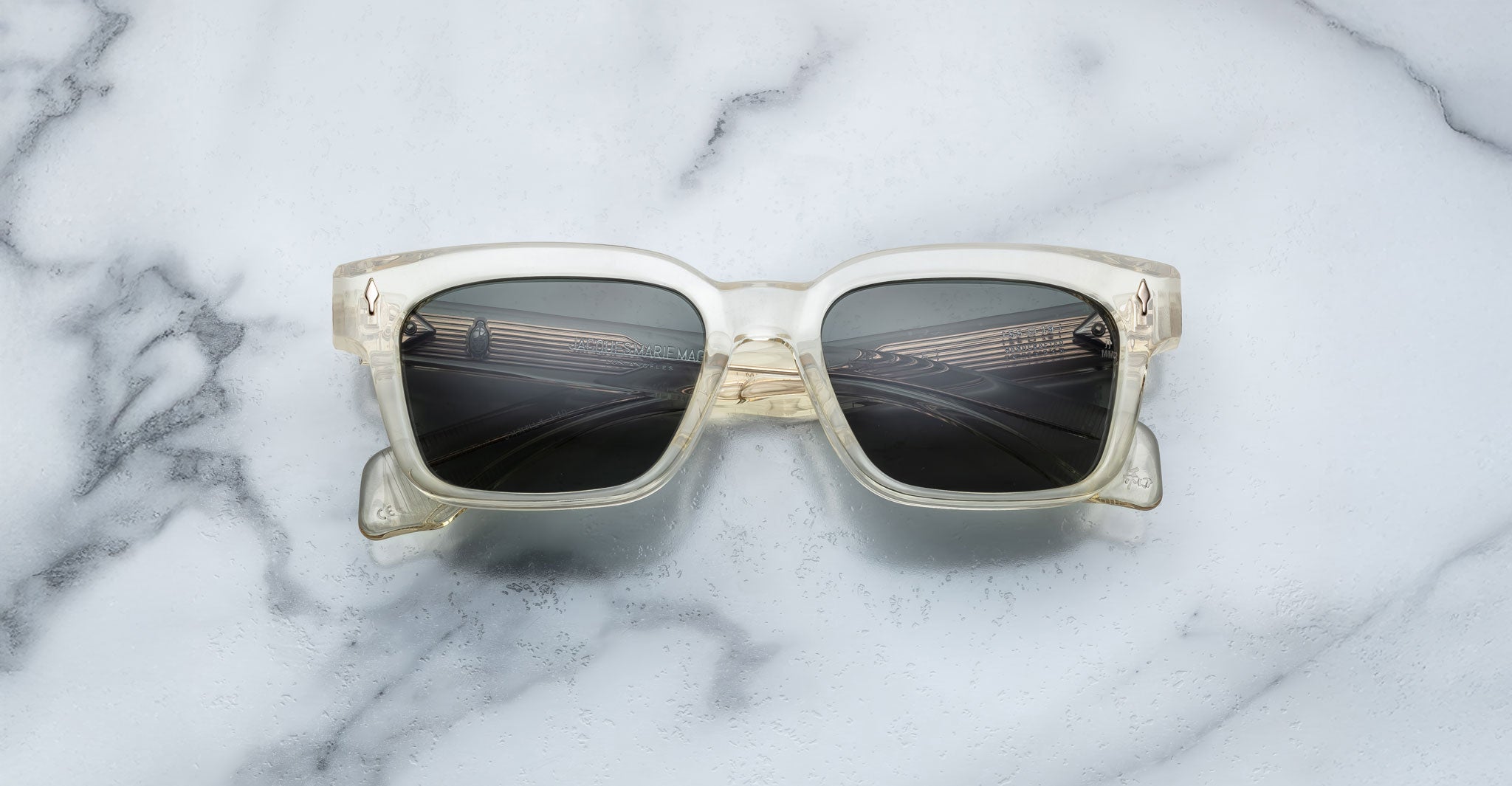 The Jacques Marie Mage Molino 55 sunglasses with clear square frames and dark lenses are centered on a white marble surface with gray veins, casting a faint shadow.