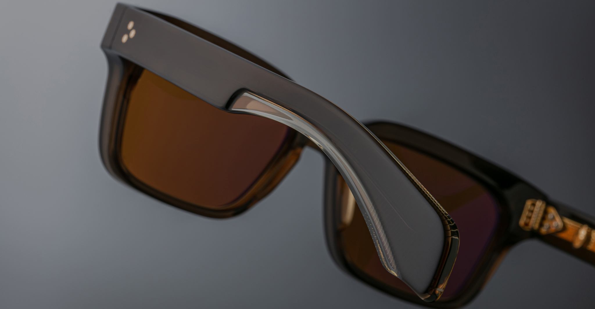 Close-up of Jacques Marie Mage Molino 55 sunglasses featuring thick dark brown frames and amber-tinted lenses, with focus on the arm and hinge, set against a blurred gray background.