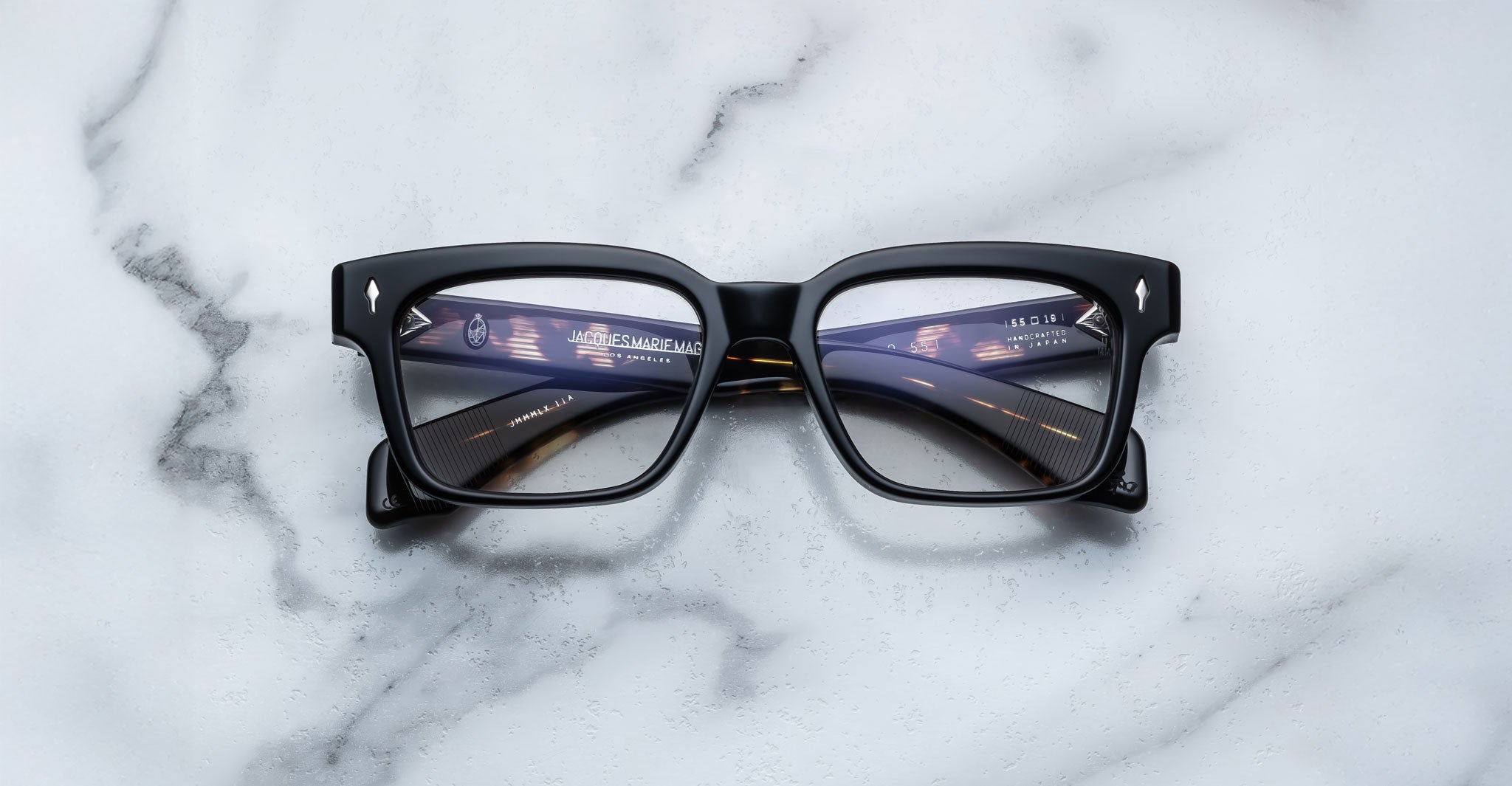 A pair of Jacques Marie Mage Molino 55 black rectangular eyeglasses rests on a white marble surface, with digital tunnel and interface graphics reflected on the lenses.