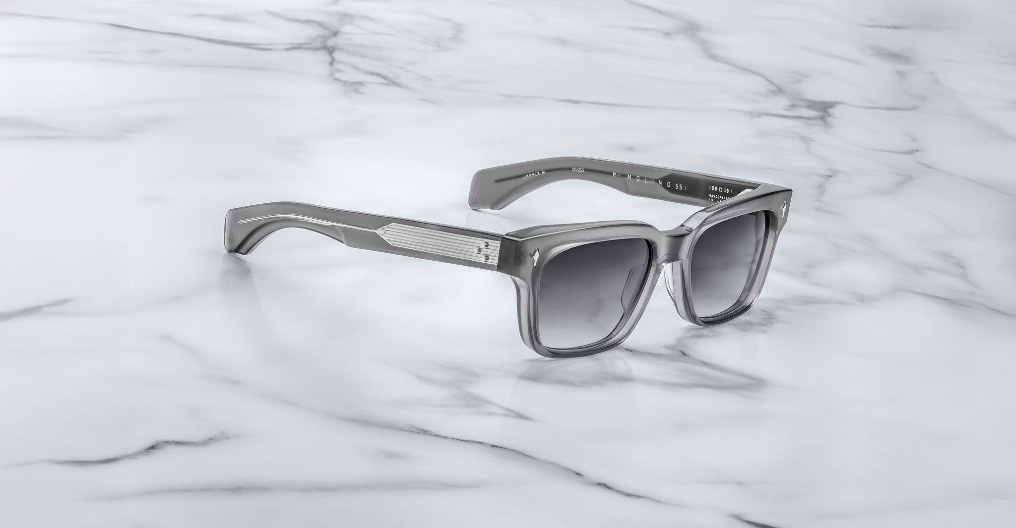 A pair of Jacques Marie Mage Molino 55 gray square-frame sunglasses with dark lenses rests on a white marble surface.