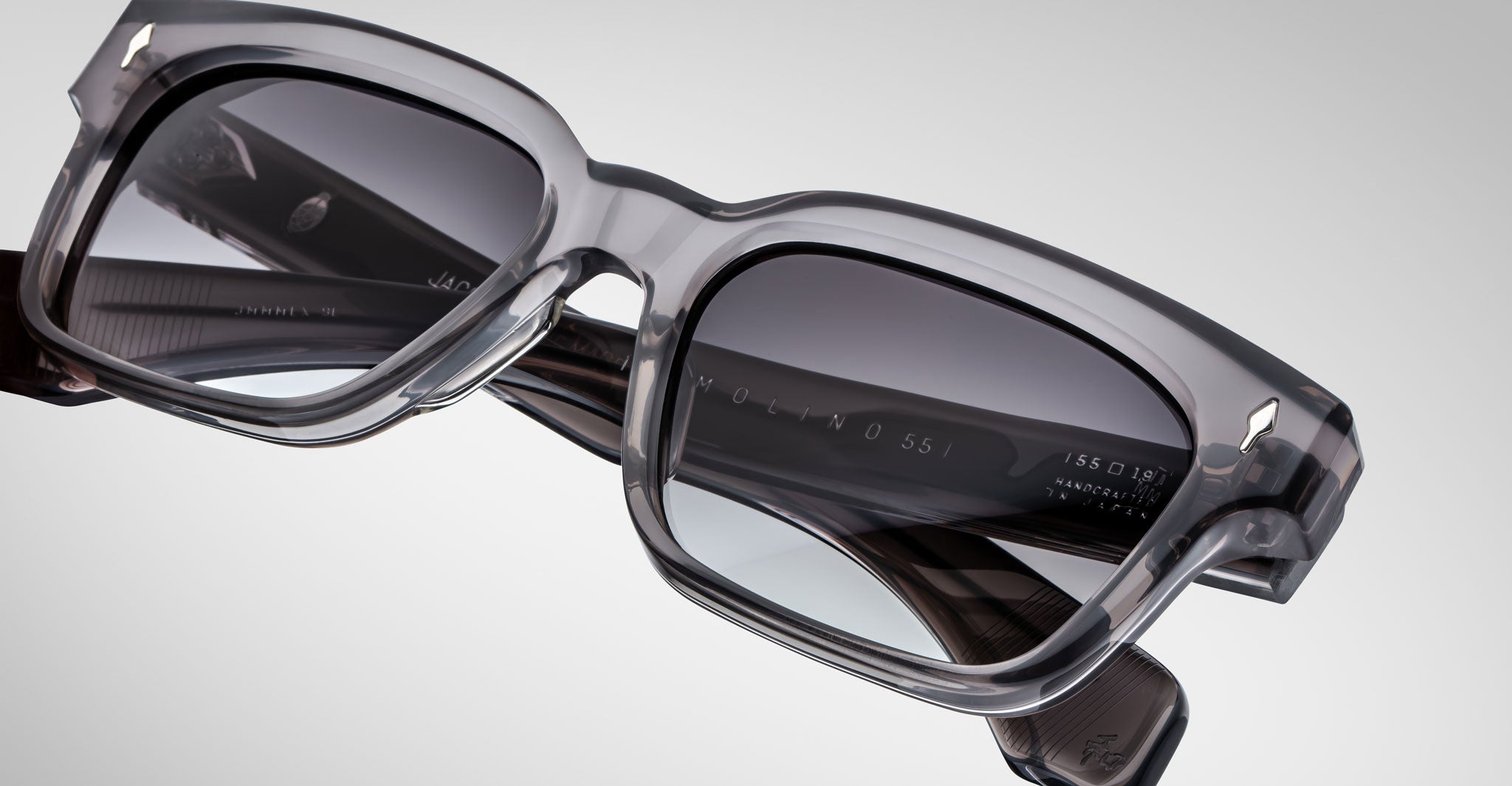 Close-up of Jacques Marie Mage Molino 55 sunglasses—stylish, rectangular gray eyewear with thick, transparent frames and dark-tinted lenses set against a plain, light background.