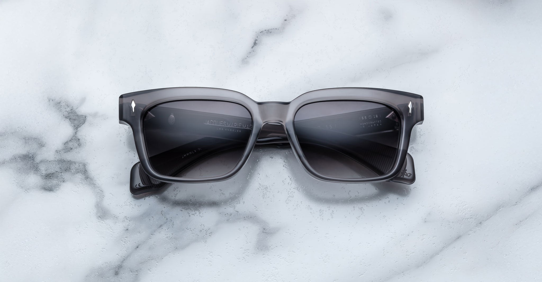 A pair of Jacques Marie Mage Molino 55 black rectangular sunglasses with dark lenses rests on a white marble surface with gray veining.
