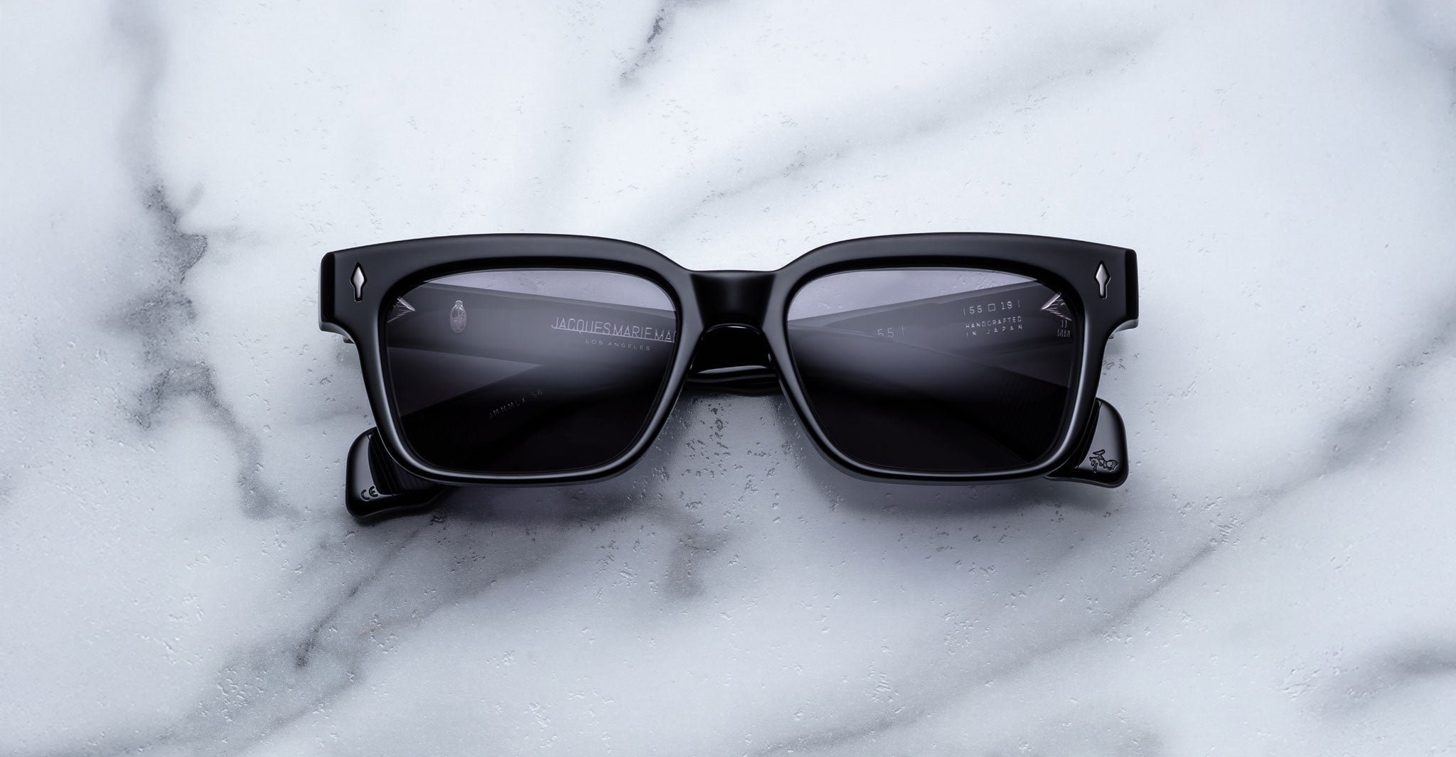A pair of Jacques Marie Mage Molino 55 black rectangular sunglasses with dark lenses rests on a white marble surface with grey veining.