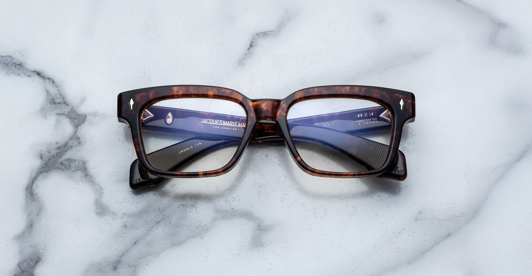 The Jacques Marie Mage Molino 55, a pair of rectangular tortoiseshell eyeglasses with clear lenses, rests on a white marble surface with gray veining.