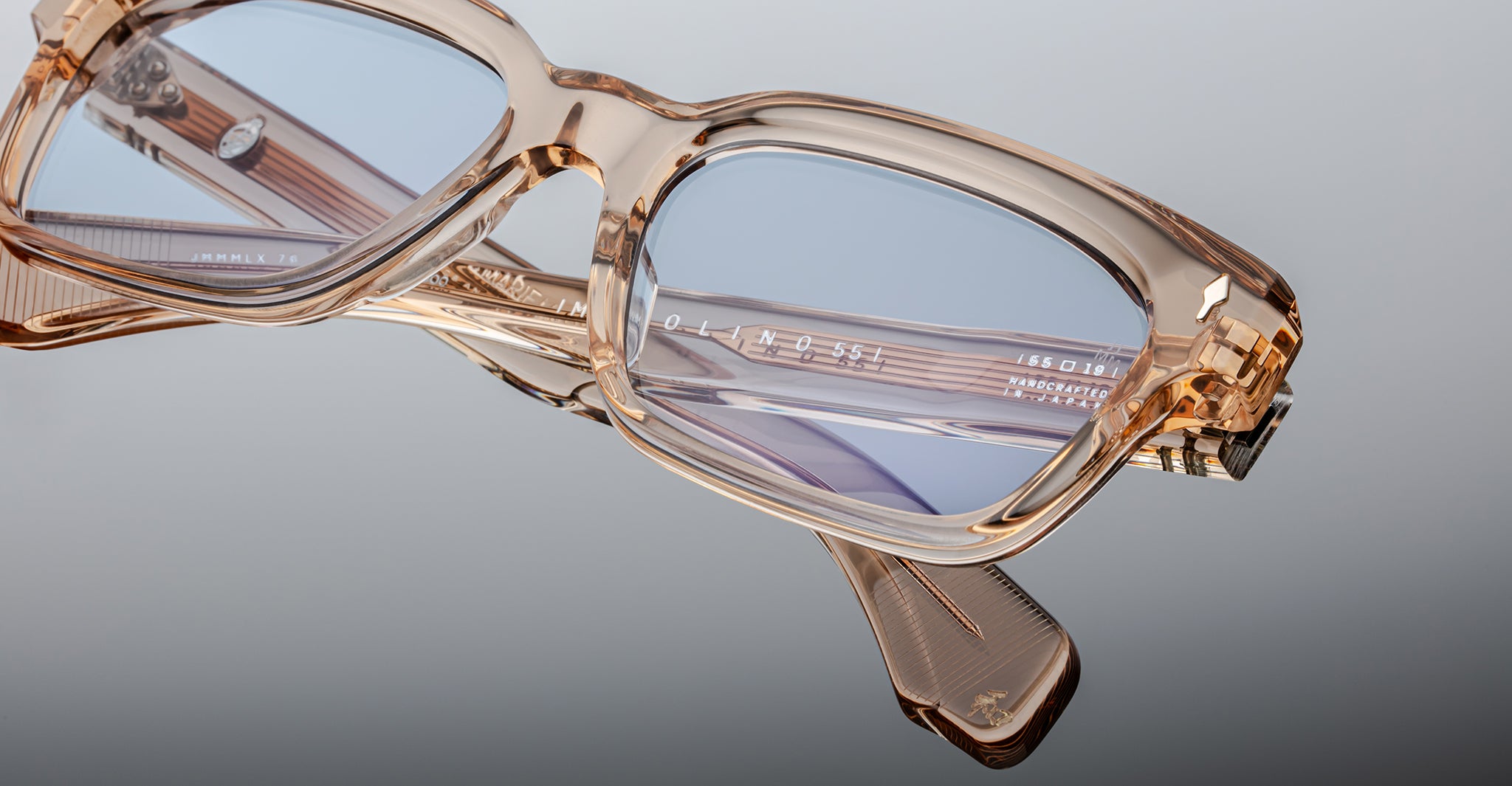Close-up of Jacques Marie Mage Molino 55 eyeglasses in transparent beige with blue-tinted lenses, featuring detailed frame design and branding on the temple arm, set against a gray background.