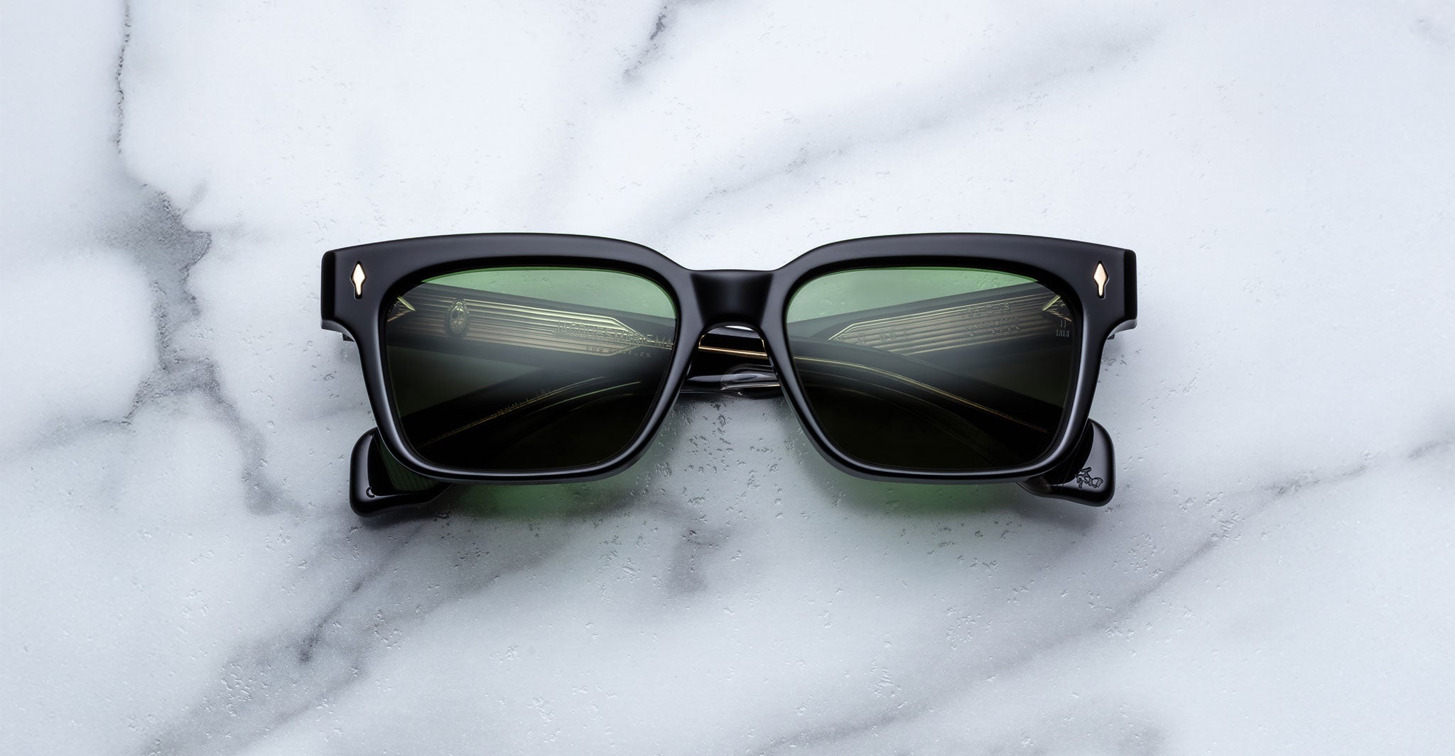The Jacques Marie Mage Molino 55, black square-framed sunglasses with green-tinted lenses, is displayed on a white marble surface. The lenses reflect a metallic or architectural structure.