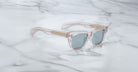 The Jacques Marie Mage Molino sunglasses by COMING SOON feature a clear, square-shaped frame with gold accents and blue-tinted lenses, displayed on a white marble surface.