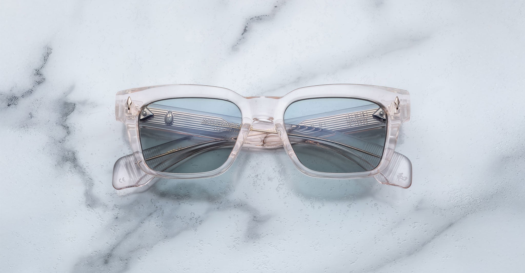 The Jacques Marie Mage Molino features square, clear frames with reflective blue-tinted lenses, shown on a white marble surface mirroring a curved metallic structure.
