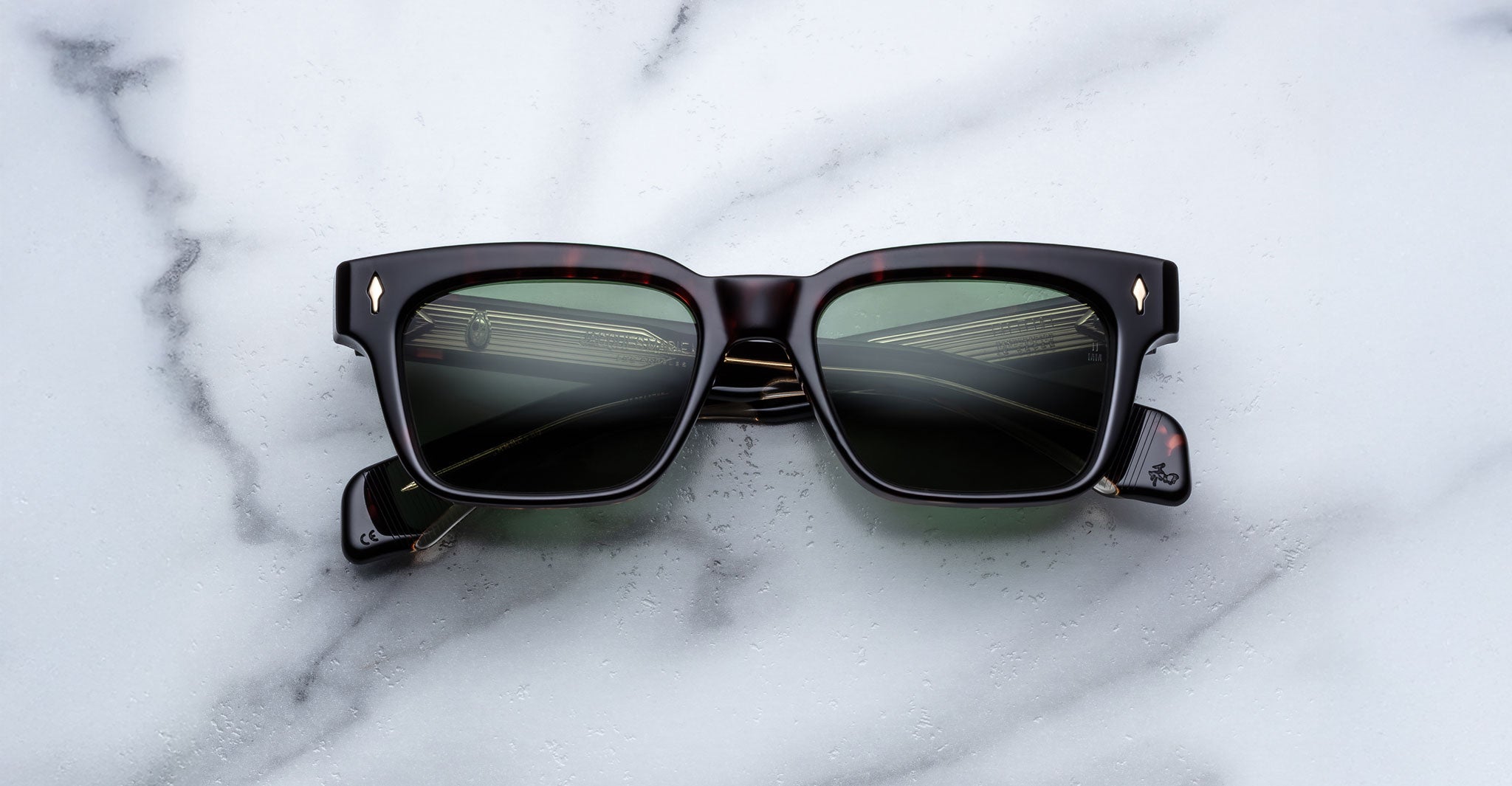 A pair of Jacques Marie Mage Molino black rectangular sunglasses with green-tinted lenses lies on a white marble surface, the lenses reflecting part of a building with windows and railings.
