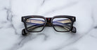 A pair of Jacques Marie Mage Molino rectangular eyeglasses with dark, patterned frames rests on a white marble surface. The temples feature gold and blue detailing visible through the lenses.