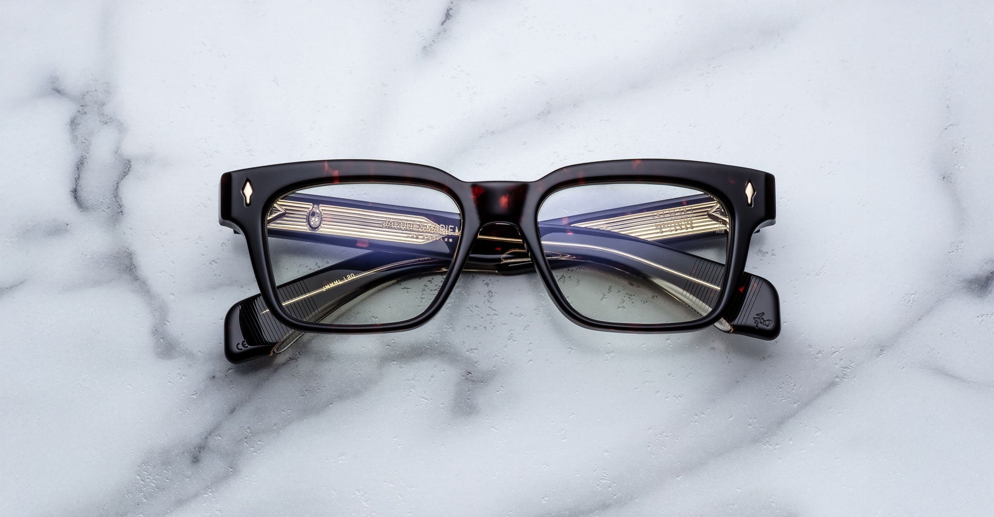 A pair of Jacques Marie Mage Molino rectangular eyeglasses with dark, patterned frames rests on a white marble surface. The temples feature gold and blue detailing visible through the lenses.