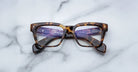 A pair of Jacques Marie Mage Molino rectangular tortoiseshell eyeglasses with clear lenses rests on a white marble surface, showcasing text and branding on the inner arms visible through the lenses.