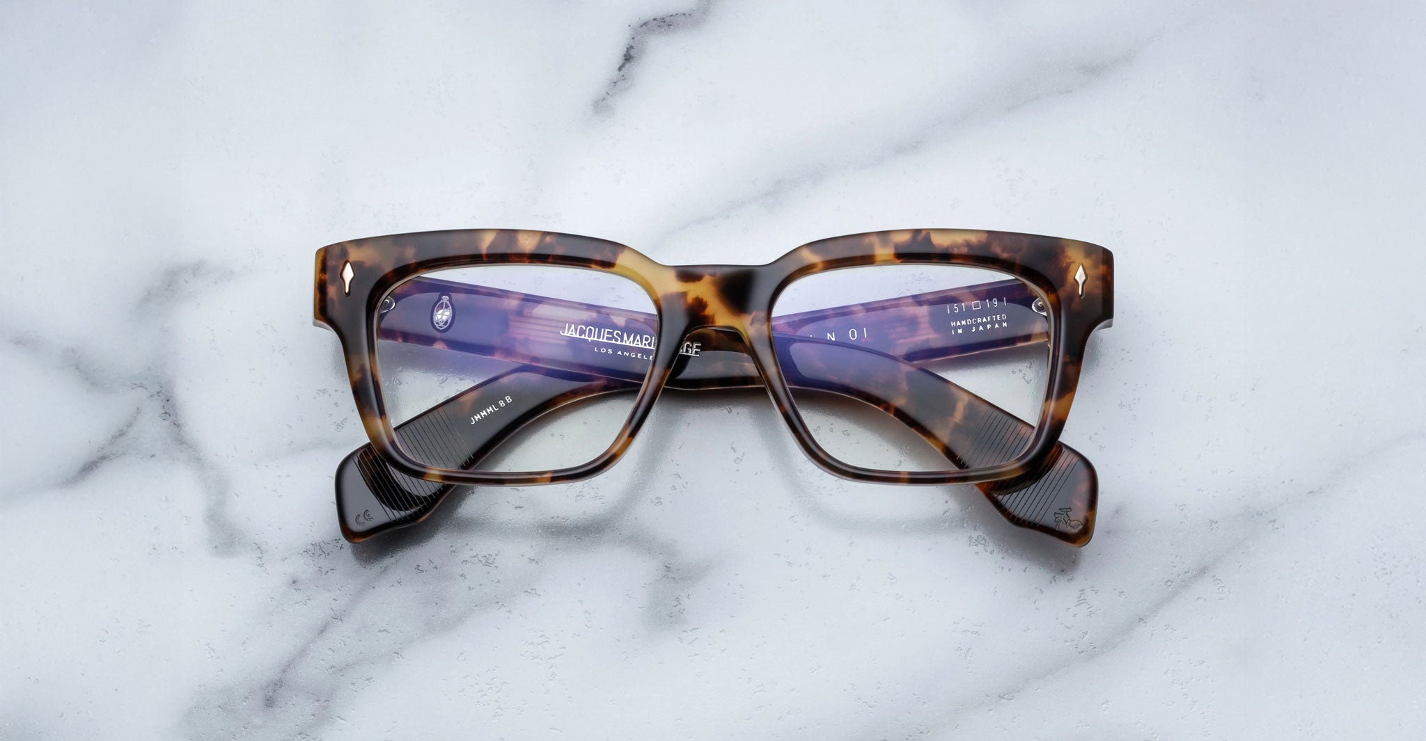 A pair of Jacques Marie Mage Molino rectangular tortoiseshell eyeglasses with clear lenses rests on a white marble surface, showcasing text and branding on the inner arms visible through the lenses.