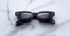 A pair of Jacques Marie Mage Molino black rectangular sunglasses with dark lenses rests on white marble, featuring silver corner accents and subtle branding on the temples.