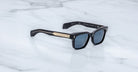 The Jacques Marie Mage Molino sunglasses feature black frames, dark lenses, and gold metallic temple accents, displayed on a white marble surface.