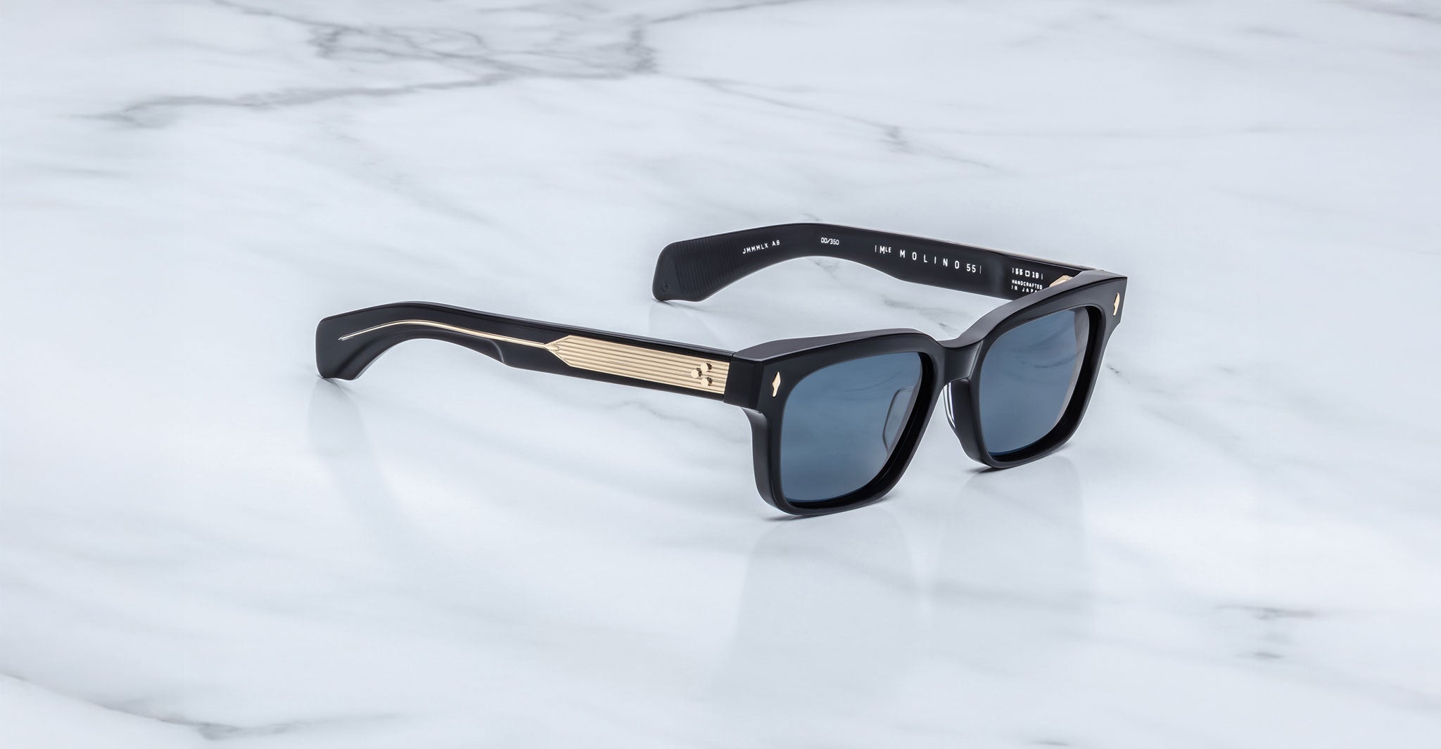 The Jacques Marie Mage Molino sunglasses feature black frames, dark lenses, and gold metallic temple accents, displayed on a white marble surface.