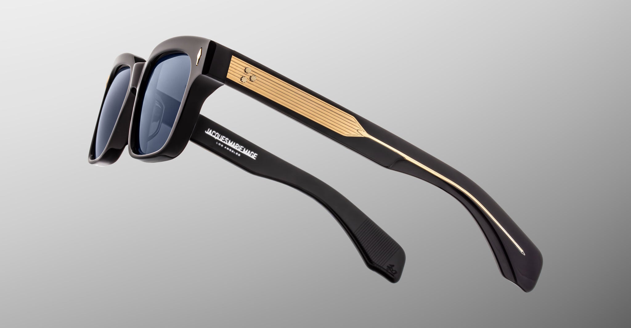 The Jacques Marie Mage Molino by COMING SOON features black sunglasses with dark lenses, thick arms, gold detailing on the sides, and a metallic accent near the hinges, displayed against a gradient gray background.