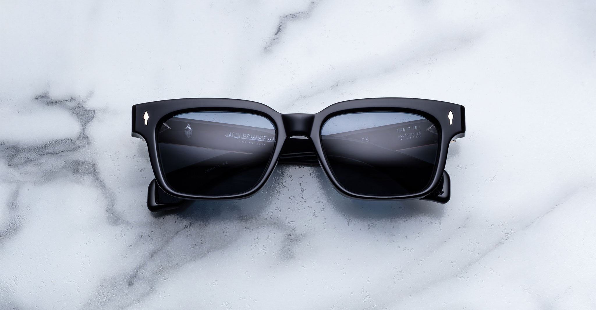 The Jacques Marie Mage Molino a pair of black square sunglasses with dark lenses, rests on a white marble surface, reflecting light and faint text in the lenses.