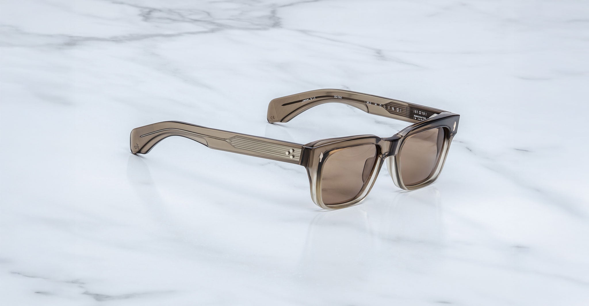 The Jacques Marie Mage Molino sunglasses feature square, brown-tinted lenses and thick, translucent brown frames, shown resting on a white marble surface.