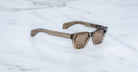 The Jacques Marie Mage Molino sunglasses feature square, brown-tinted lenses and thick, translucent brown frames, shown resting on a white marble surface.
