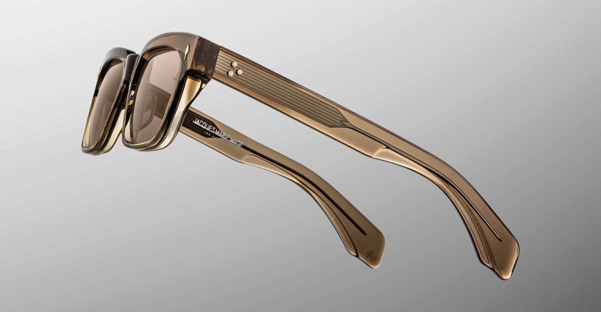 A close-up of Jacques Marie Mage Molino sunglasses by COMING SOON, featuring translucent brown frames, thick textured arms, and light brown tinted lenses, set against a neutral gray gradient background.