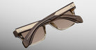 The Jacques Marie Mage Molino by COMING SOON are stylish, square-shaped brown sunglasses with gold accents and ribbed temple detailing, shown folded on a light gray background.
