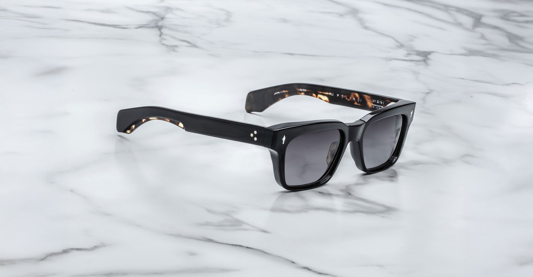 The Jacques Marie Mage Molino by COMING SOON features thick, glossy black rectangular frames with dark lenses and small silver corner accents, displayed on a white marble surface.