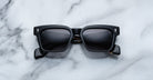 Jacques Marie Mage Molino by COMING SOON are black rectangular sunglasses with thick frames and dark lenses, resting on a white marble surface. The curved arms feature a subtle design detail near the hinges.