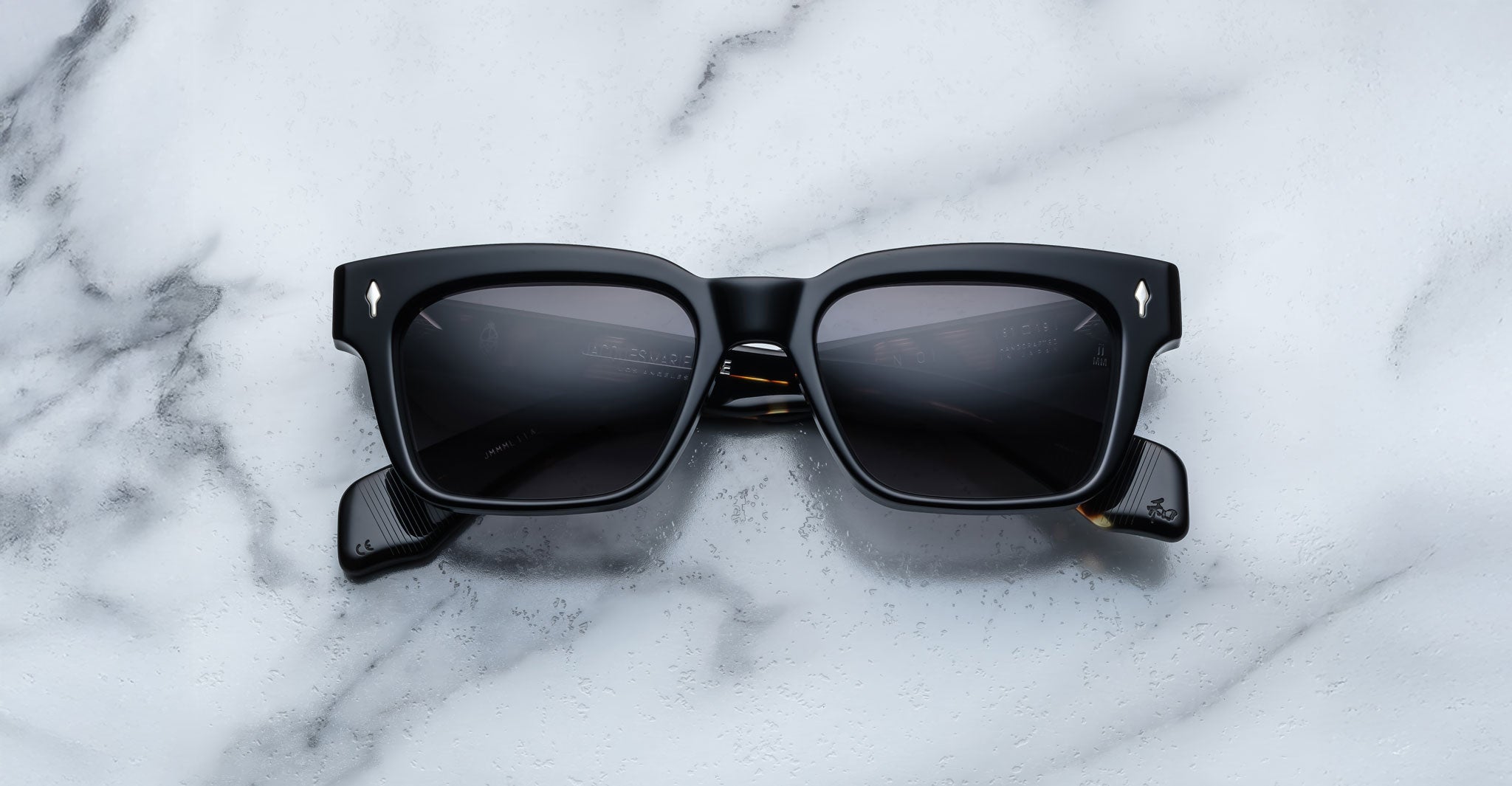 Jacques Marie Mage Molino by COMING SOON are black rectangular sunglasses with thick frames and dark lenses, resting on a white marble surface. The curved arms feature a subtle design detail near the hinges.