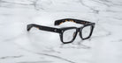 Jacques Marie Mage Molino black rectangular eyeglasses with thick frames displayed on a white marble surface with grey veining.