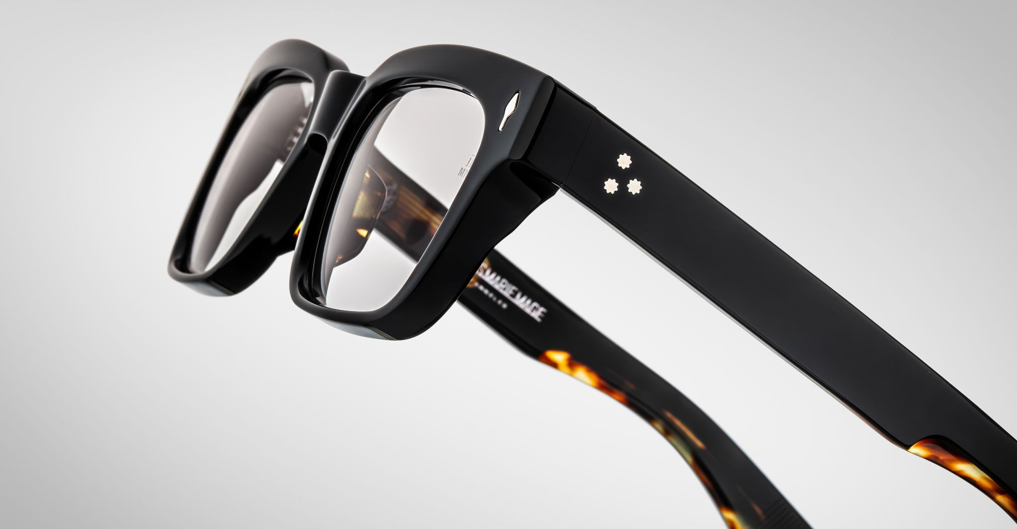 Close-up of the Jacques Marie Mage Molino by Jacques Marie Mage, showcasing stylish thick black frames with silver dot accents on the arms and tortoiseshell inner details, set against a light gradient background.