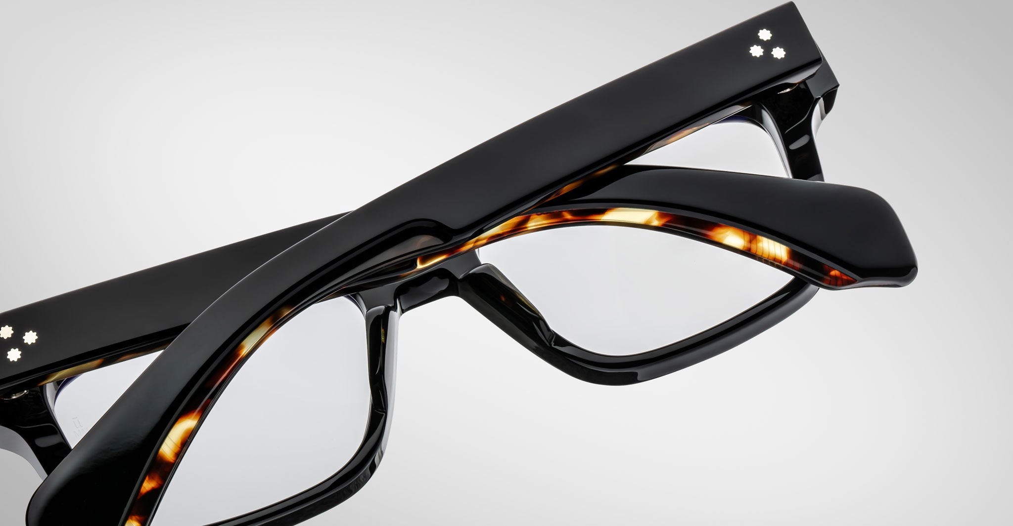 Stacked are two pairs of Jacques Marie Mage Molino by COMING SOON—one in glossy black, the other in tortoiseshell—each with clear lenses and silver decorative dots on the temples.
