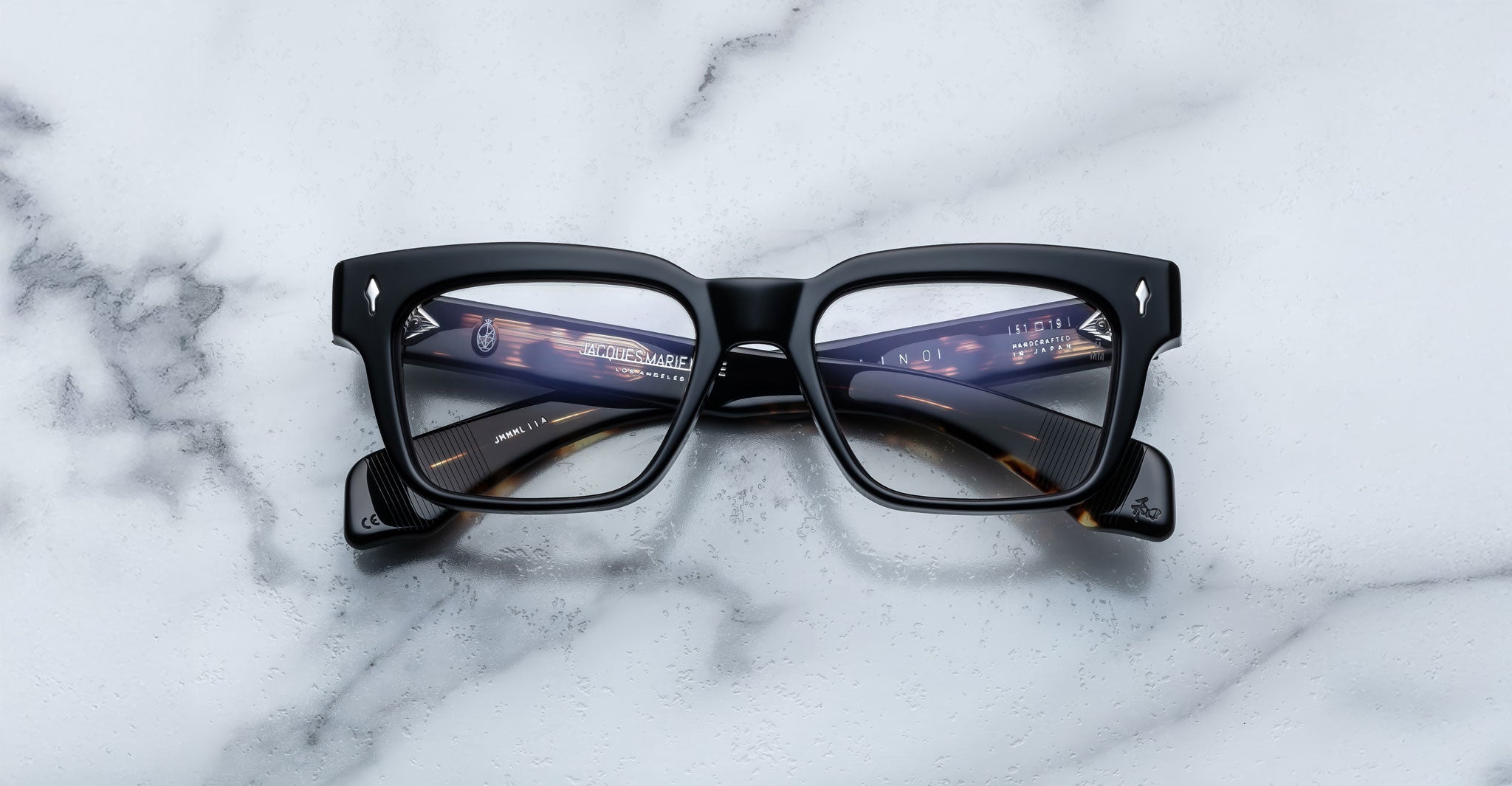 A pair of Jacques Marie Mage Molino eyeglasses rests on a white marble surface, with their lenses reflecting a futuristic cityscape filled with bright lights and digital elements.