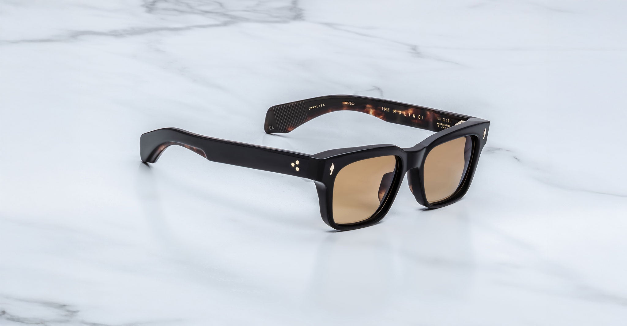 The Jacques Marie Mage Molino by COMING SOON, featuring rectangular dark frames and brown-tinted lenses, rests on a white marble surface.